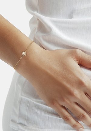 Fijne gouden kettingarmband met een klein hartvormig bedeltje, gedragen om de pols tegen een witte textielachtergrond.