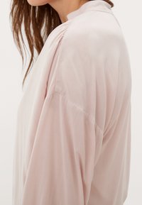 Blouse rose clair à texture lisse, avec des manches longues et un col montant. Un ruché à l'arrière ajoute un détail subtil au design.