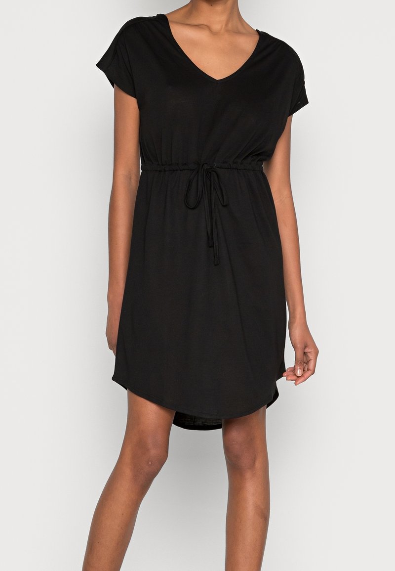 Femme portant une robe courte noire avec un col en V et une taille à cordon, debout devant un fond uni gris clair.