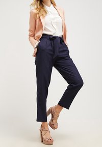 Námořnické kalhoty na míru mají vysoký pas a látkový pásek, kombinují se s bílou košilí a světle růžovým blejzrem, doplněny béžovými platformovými sandály.