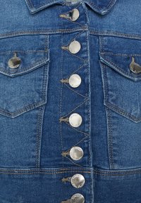 Denim jakna v srednje modri barvi z srebrnimi zapirali, dvema žepoma na prsih in kontrastnim šivanjem, ki ima klasičen ovratnik in teksturirano blago.