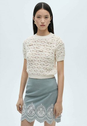 Gestrickter cremefarbener Pullover mit kurzen Ärmeln, der ein spitzenartiges Design und florale Akzente aufweist, kombiniert mit einem mintgrünen Rock, der mit aufwendiger Spitzenborte verziert ist.