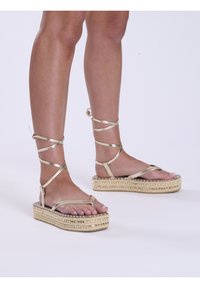 Sandali dorati con cinturini, dal design flat, caratterizzati da una base piatta intrecciata e dettagli testurizzati lungo i bordi.