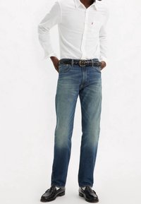 Aansluitende blauwe denim jeans met een lichte vervaging, aangevuld met een zwarte gevlochten riem. Het model draagt een wit shirt met lange mouwen en zwarte loafers.