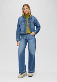 Blaue Jeansjacke mit zwei Brusttaschen, kombiniert mit einem grünen Rollkragenpullover. Breite blaue Jeans und beige Plattformschuhe sind ebenfalls zu sehen.