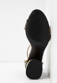 Semelle d’une sandale à talons hauts avec semelle extérieure noire et brides à imprimé léopard, positionnée à la verticale contre un coin blanc.