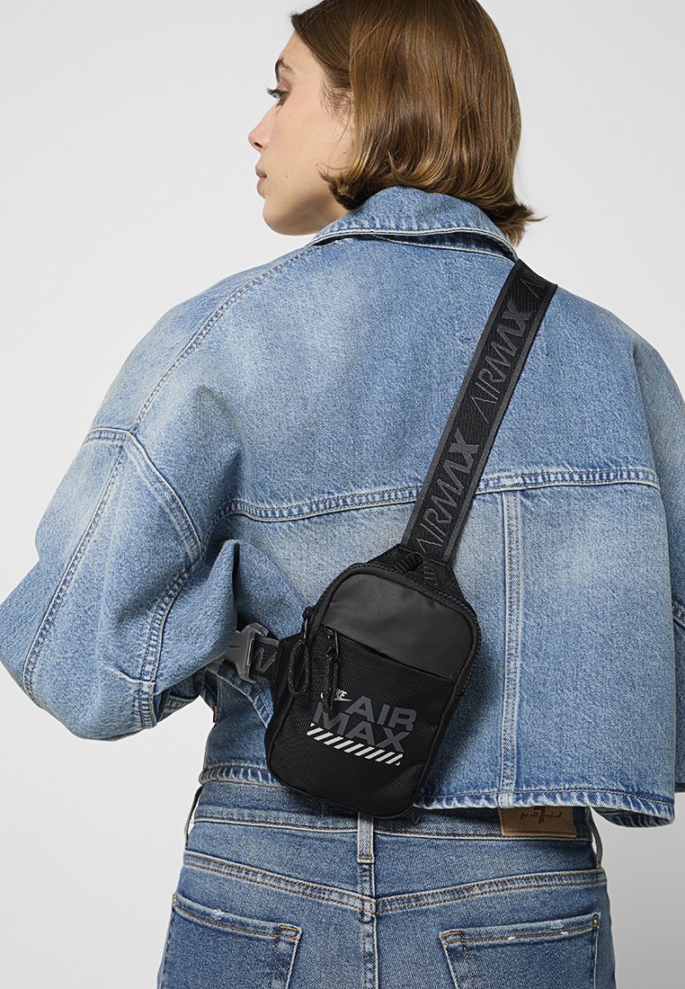 Bolso cruzado de nylon negro con texto reflectante "AIR MAX", correa ajustable y superficie texturizada. Lleva puesto sobre una chaqueta de denim azul.