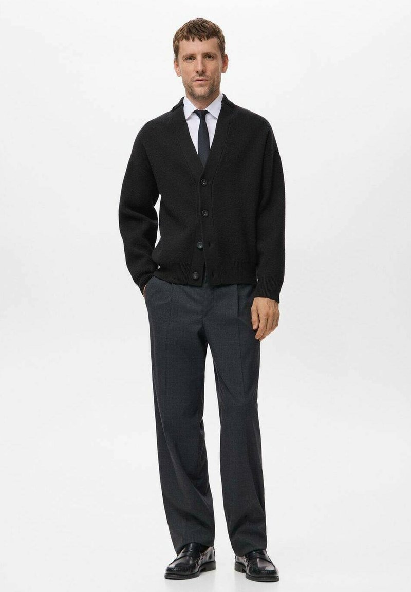 Zwarte gebreide cardigan met een V-hals, zes knopen en geribbelde manchetten. Combinatie met grijze broeken en zwarte leren schoenen. Effen achtergrond.