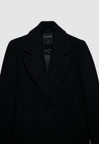 Cappotto di lana nera con ampio colletto a rever, chiusura con un bottone e due tasche anteriori. Presenta una foderatura interna liscia. Taglia: S.