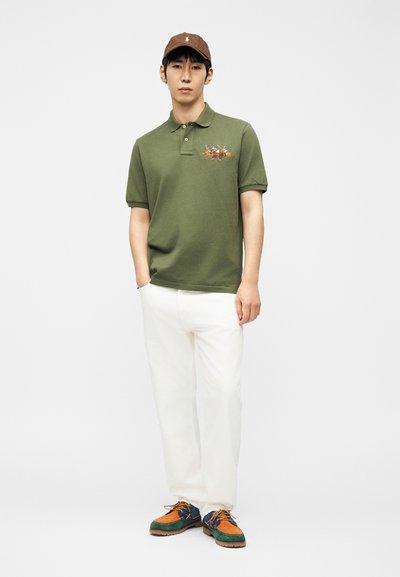 Άνδρας που φορά καφέ καπέλο, πράσινο πόλο με κέντημα παίκτη πόλο με άλογο, λευκό παντελόνι και πολύχρωμα καθημερινά παπούτσια, στέκεται με τα χέρια στις τσέπες.