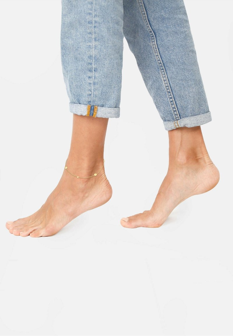 Pieds nus avec une cheville en or levée à mi-pas, portant un jean en denim bleu clair retroussé sur fond blanc.