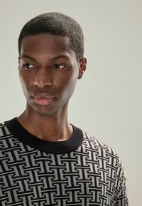 Pulover cu model alb și negru, cu decolteu rotund. Prezintă un design geometric și un material texturat. Prim-plan al feței unui model masculin.