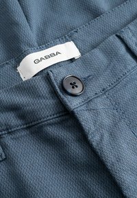 Mėlynos austos medžiagos kelnės su sagos uždarymu ir etikete, kurioje parašyta „GABBA“. Tekstūruotas paviršius su subtiliu raštu.