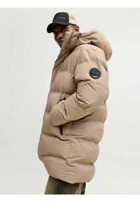 Chaqueta acolchada beige con cuello alto y capucha. Presenta un logotipo negro redondo en la manga izquierda y bolsillos laterales con cremallera.