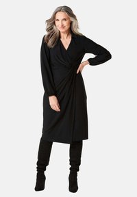 Schwarzes Wickelkleid mit tiefem V-Ausschnitt, langen Ärmeln und gerafften Details an der Taille. Weicher Stoff kombiniert mit schwarzen kniehohen Stiefeln.