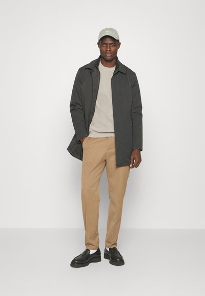 Chaqueta gris oscuro, suéter beige, pantalones color crema, zapatos negros y una gorra verde claro. El modelo está de pie contra un fondo blanco.