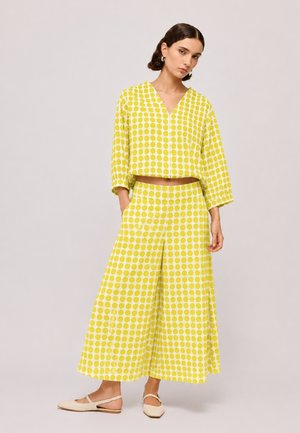 Vrouw draagt een bijpassende geel-witte polkadot top met lange mouwen en wijd uitlopend cropped broek met beige platte schoenen, staand tegen een effen achtergrond.