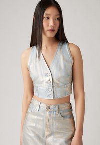 Metallic denim cropped vest in light blue with silver accents, featuring a v-neckline and front buttons. Smooth texture, fitted design.
