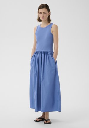 Mladá žena stojaca v modrých bezrukávových maxi šatách s vreckami a čiernymi sandálmi, neutrálne pozadie.