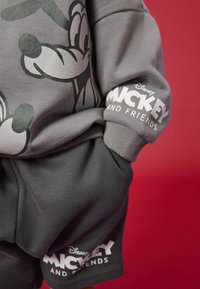 Harmaa huppari, jossa on suuri musta hahmosuunnittelu ja valkoinen "Mickey and Friends" -teksti hihassa, yhdistettynä tummansiin shortsien kanssa.