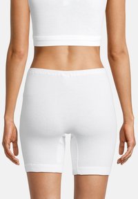 Kvinde set bagfra iført hvide tætsiddende biker-shorts og en hvid cropped tanktop mod en helt hvid baggrund.