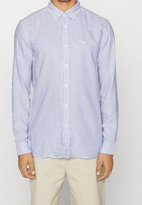 Blå och vit randig långärmad skjorta med krage, knapplist fram och en liten logotyp på bröstet. Slät tyg och standardpassform.