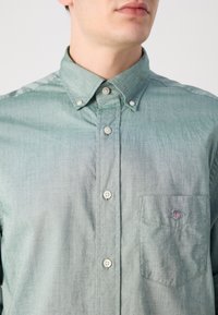 Lys grøn skjorte med knapper og brystlomme, hvite knapper og logo på lommen. Teksturert stoff med en subtil gradient.