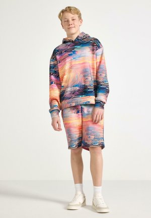 Teini-ikäinen poika seisoo, yllään värikkäällä abstraktikuvioisella hupparilla ja shortseilla, valkoiset sukat ja lenkkarit, yksivärinen tausta.