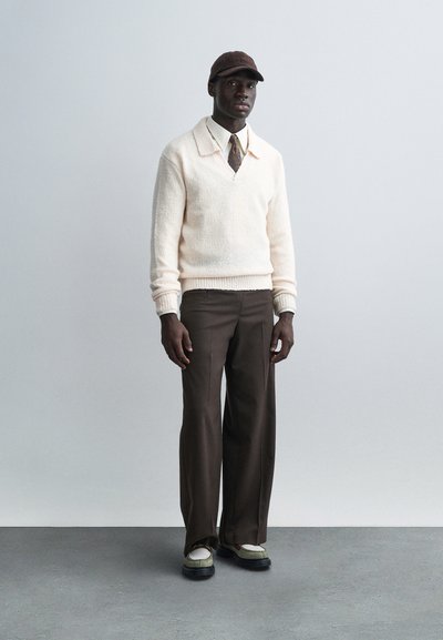 Pull en tricot crème avec col en V, pantalon marron à jambes larges, et casquette marron. Les chaussures présentent des accents blancs et noirs. Design simple et moderne.