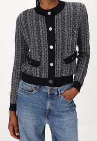 Stickad kofta med ett texturerat svart och vitt mönster, rund krage, silverfärgade knappstängningar och två sidofickor. Ribbad mudd och nederkant.