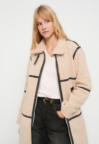 Cappotto in pile beige con accenti neri, zip frontale e ampio colletto. Indossato sopra un maglione rosa chiaro a V e jeans scuri, con cintura.