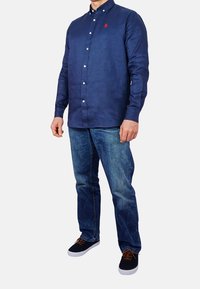 Mörkblå skjorta med knappar i linneblandning, med en liten röd logotyp på bröstet, ihop med blå jeans och svarta sneakers.