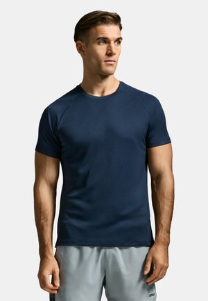 Uomo con capelli castani corti che indossa una T-shirt blu navy aderente e pantaloncini grigi chiari, che guarda leggermente verso destra.