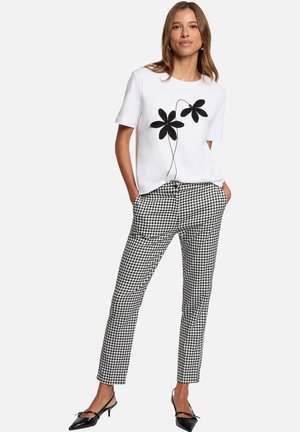 Femme portant un haut blanc à manches courtes avec un motif floral noir, un pantalon à carreaux noir et blanc, et des chaussures noires à talon bas, mains dans les poches.