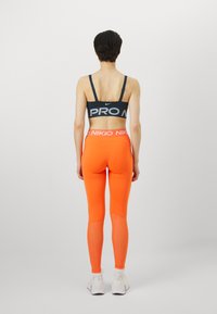 Svart sports-bh med "PRO" logo, kombinert med lyse oransje tights med bred midjebånd med "NIKKO" tekst og hvite joggesko.
