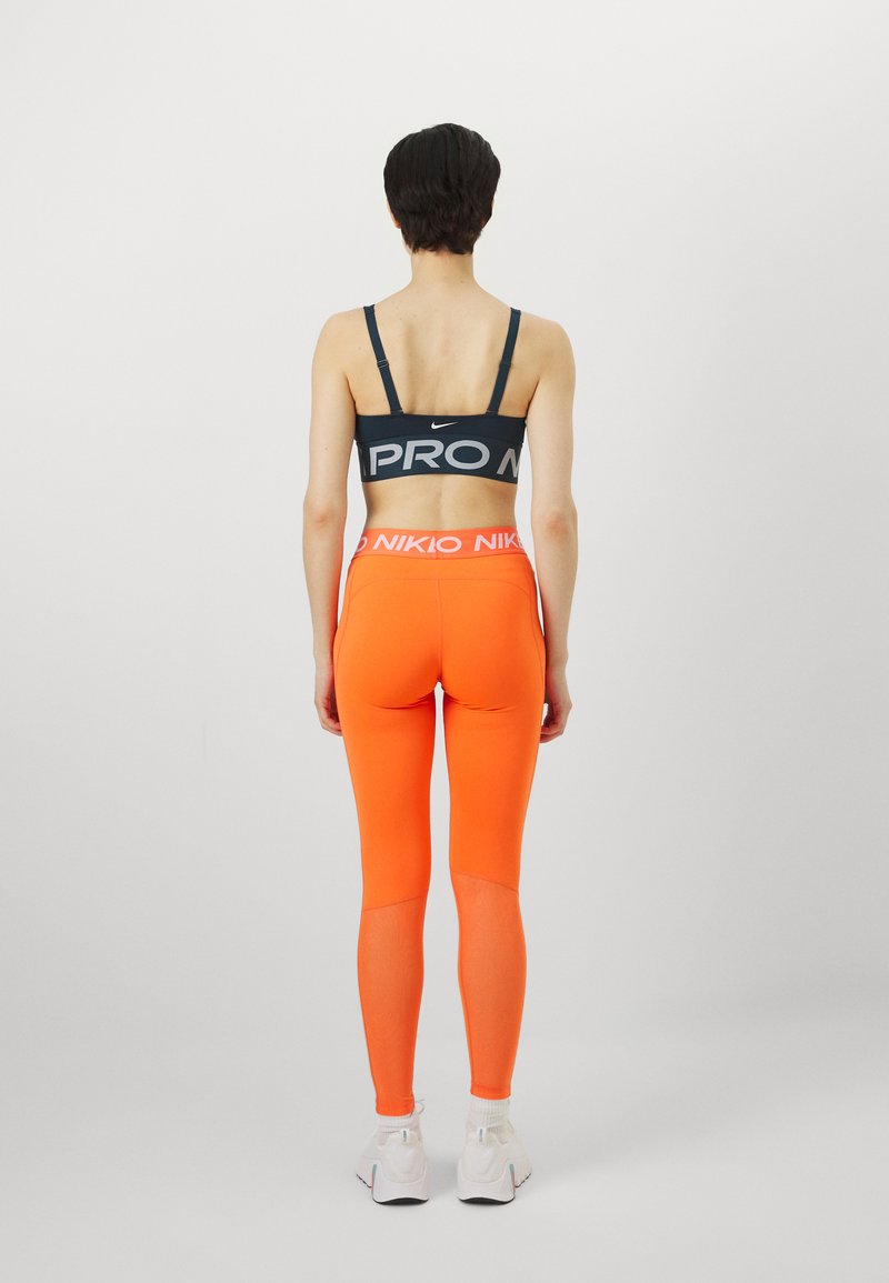 Schwarzer Sport-BH mit "PRO"-Logo, kombiniert mit leuchtend orangefarbenen Leggings, die einen breiten Bund mit "NIKKO"-Text und weißen Sneakers haben.