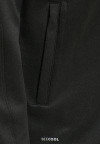 Veste de sport noire avec une texture lisse, des poches latérales et un petit logo blanc en bas à droite.