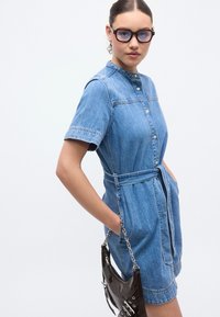 Φόρεμα από τζιν ύφασμα με κοντά μανίκια, κουμπιά μπροστά, ζωνάκι στη μέση και πλαϊνές τσέπες. Το μοντέλο κρατάει μια λαμπερή μαύρη τσάντα χειρός με αλυσίδα.