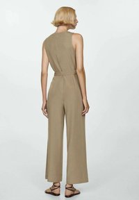 Kvinna med kort blont hår som bär en ärmlös beige jumpsuit och bruna sandaler med remmar, sedd bakifrån mot en enfärgad bakgrund.