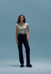 Lichtgrijze T-shirt met korte mouwen, hoge zwarte jeans en zwarte schoenen. Model staat tegen een lichtblauwe achtergrond, met golvend haar.