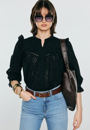 Kobieta w czarnej bluzce z ażurowym wzorem, niebieskich dżinsach, dużych okularach przeciwsłonecznych, złotych bransoletkach, niosąca ciemną skórzaną torebkę na ramię.