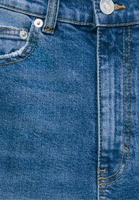 Gros plan d’un jean en denim bleu affichant la poche avant, la passants de ceinture, le bouton en métal, les rivets et les détails de couture orange.