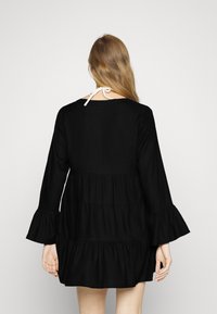 Vestido negro de capas con mangas largas y acampanadas. Presenta un escote redondo, textura fruncida y un cierre de lazo en la parte posterior.