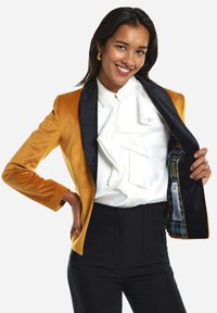 Blazer de terciopelo amarillo con solapas de satén negro, combinado con una blusa blanca con volantes y pantalones de cintura alta negros. El interior presenta forro de cuadros.