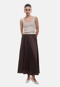 Beige geribbelde tanktop gecombineerd met een vloeiende, donkerbruine maxi rok, voorzien van een elastische tailleband en een gladde textuur. Draagt witte hakken.