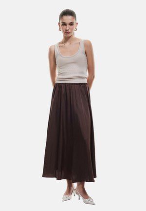 Débardeur beige à côtes associé à une longue jupe maxi fluide de couleur marron foncé, avec une taille élastique et une texture douce. Porté avec des talons blancs.