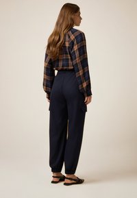 Camisa de cuadros marrón y azul marino con mangas abullonadas, combinada con pantalones azul marino con cordón, que tienen bolsillos laterales y puños elásticos, usados con sandalias negras.