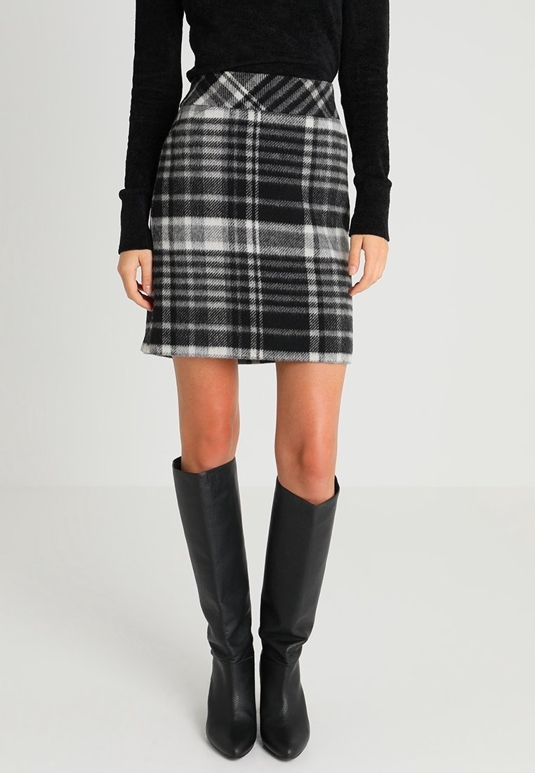 Femme portant une jupe à carreaux noire et blanche, un haut noir à manches longues et des bottes noires montantes, debout devant un fond uni.