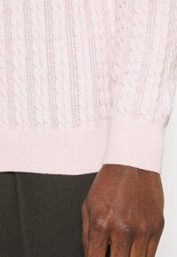 Light pink knitted sweater with ribbed cuffs, featuring a cable pattern and open stitches, paired with a dark textured fabric below.