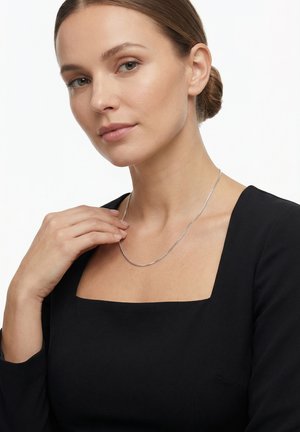 Femme à la peau claire et aux cheveux bruns attachés en chignon, portant un haut noir à col carré et un fin collier en chaîne argentée, touchant délicatement le collier.
